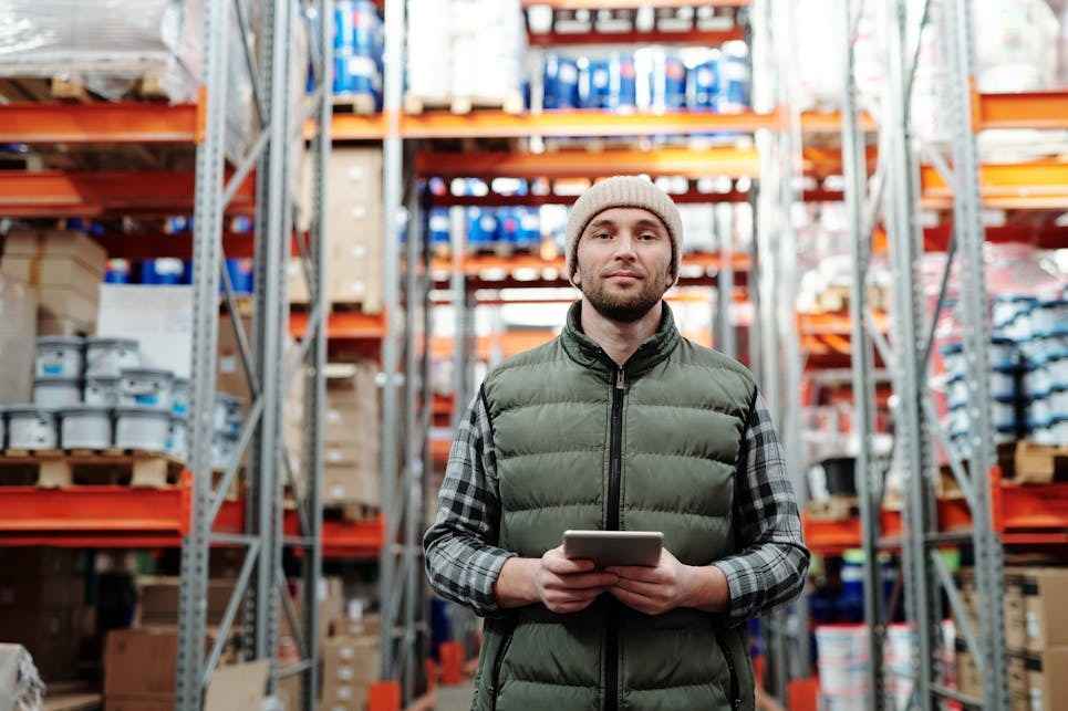 A man stands in a warehouse, holding a tablet and looking at the screen, surrounded by shelves of inventory.