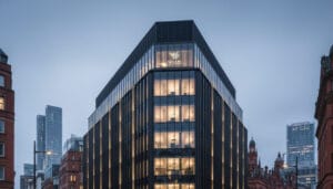 (An imposing, modern building with a dark, ribbed facade and illuminated windows, likely a law firm, stands prominently on a bustling Manchester street at dusk.)
