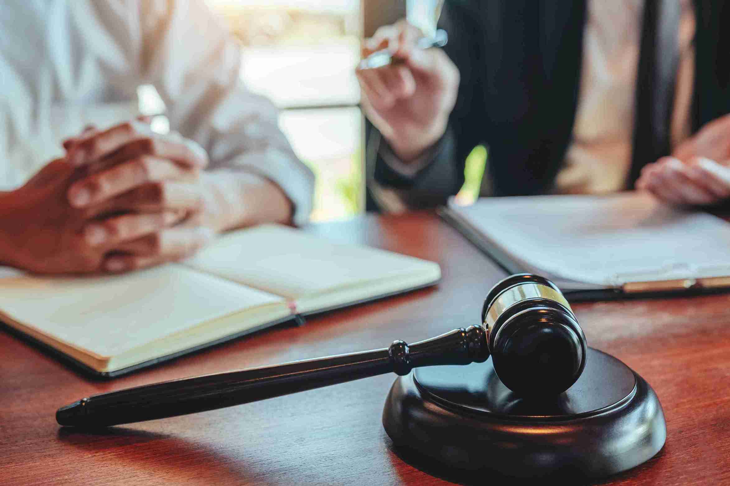 A gavel rests on a wooden table in front of two people with notebooks, suggesting a meeting or discussion related to legal matters.